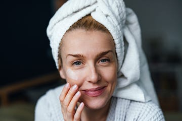 close up photo of happy woman in bathrobe using face cream and looking at camera while sitting in the bedroom