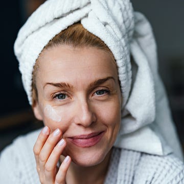 Portrait Of A Smiling Woman Applying Moisturiser Cream In The Morning close up photo of happy woman in bathrobe using face cream and looking at camera while sitting in the bedroom