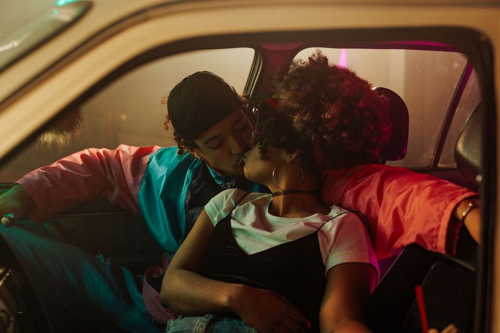 a vintage styled couple is in a car, kissing in the parking lot illuminated by neon lights