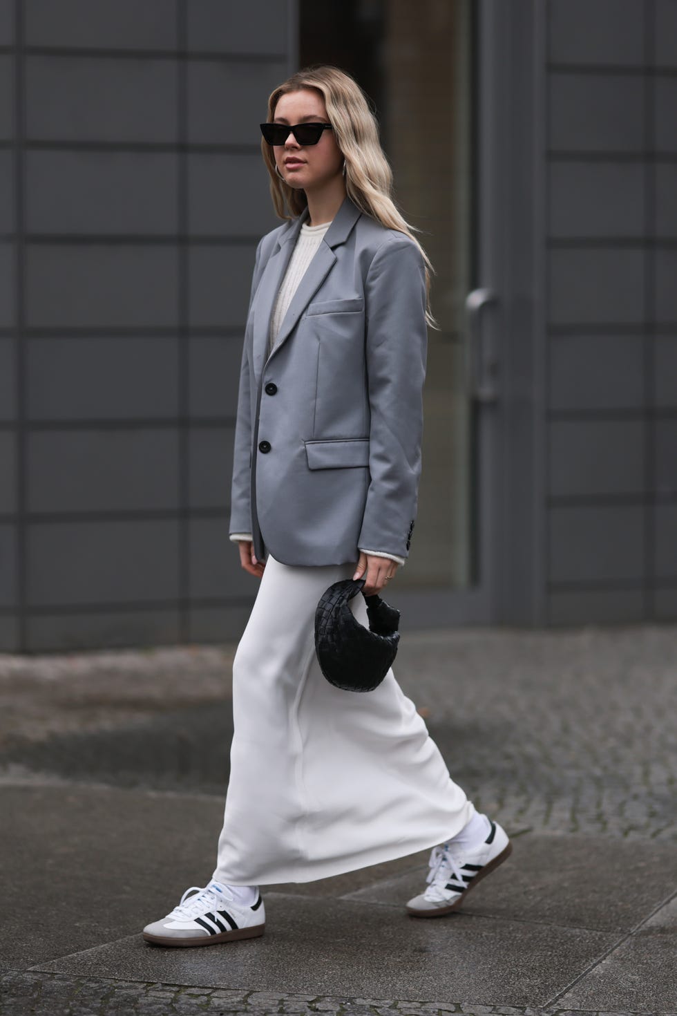 a person is seen walking on a wet pavement, dressed in a gray blazer over a white dress