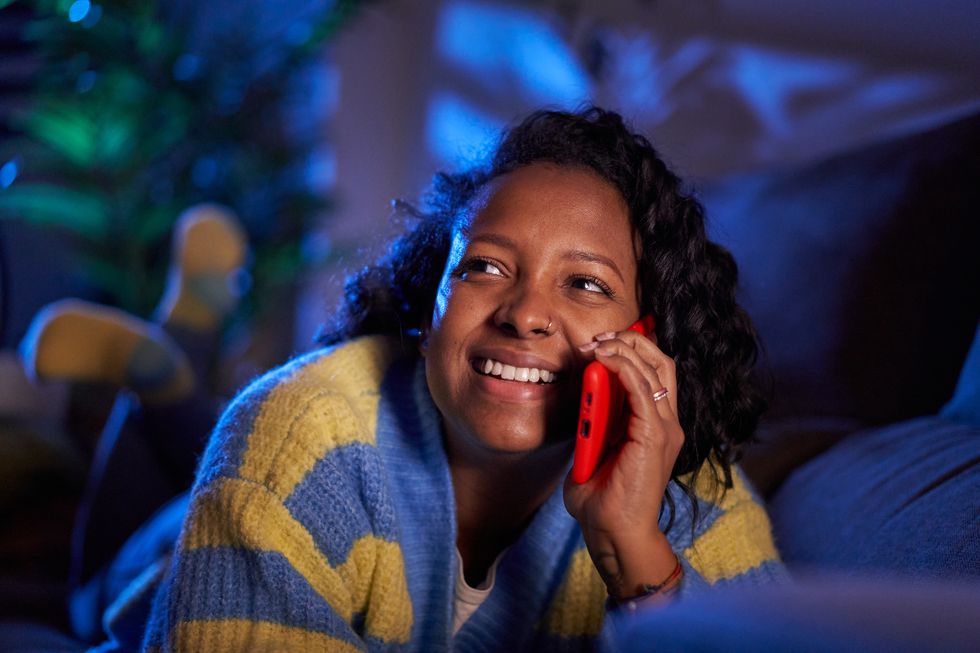 shot of a young black woman talking on a cellphone at home lying on sofa during nighttime, looking away cheerfully close up