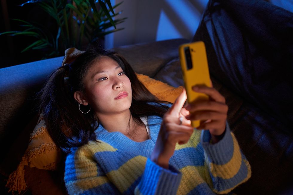 young beautiful chinese woman relaxing lying on the couch at home and texting on smartphone