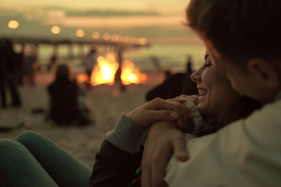 young couple sitting on the beach at sunset and laughing and hugging with campfire in background