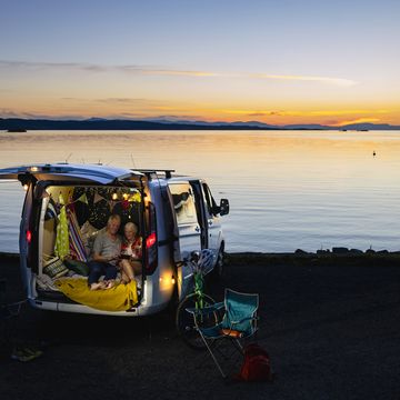 a high wide angle front view of a serene moment at sunset of a senior couple who are having a snuggle and watching something on a mobile phone in the back of their van which they have decorated with string lights and made comfortable with blankets they are on the coast in torridon in scotland