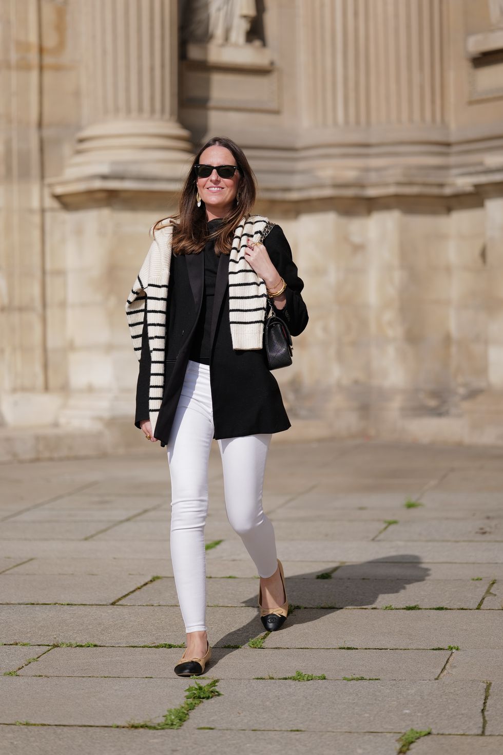 paris, france april 29 alba garavito torre wears black sunglasses from ray ban, gold and white pendant large earrings, a black t shirt, a white with black small striped print pattern wool pullover from massimo dutti, a black shiny grained leather timeless shoulder bag from chanel, a black oversized blazer jacket from munthe, white skinny denim pants from zara, beige shiny leather with black toe cap ballerinas from chanel , during a street style fashion photo session, on april 29, 2023 in paris, france photo by edward berthelotgetty images