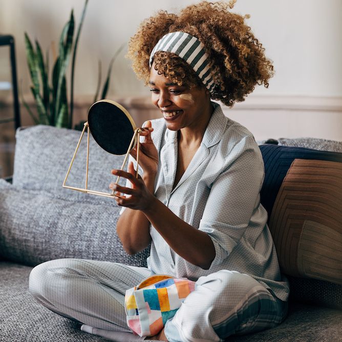 a wide angle view of a smiling african american female holding a mirror while applying some creme on her face to keep it fresh and hydrated she is sitting on the sofa in the living room skincare concept