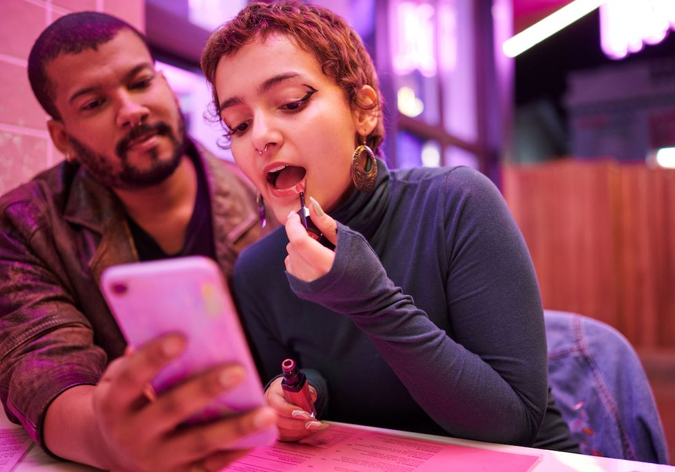 man using a phone screen to help his friend put on lipstick while sitting at a table in a bar during night out