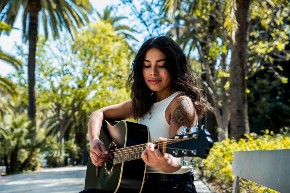 portrait of a south american woman with tattoos sitting on a bench in a park playing her guitar.
