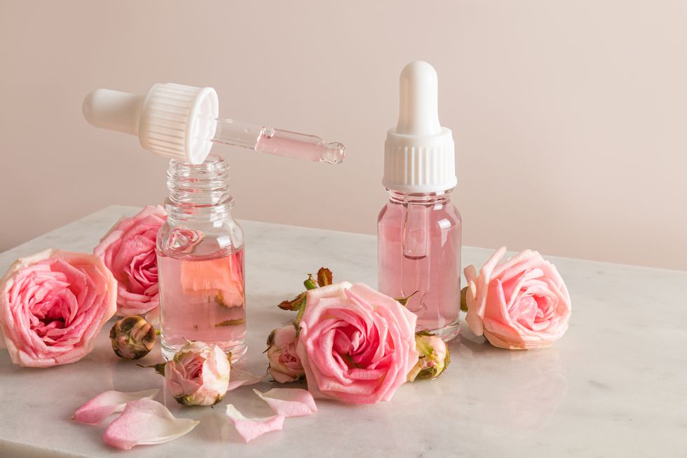 an open bottle of rose oil and a pipette filled with cosmetics on a marble podium with flowers and rose petals moisturizing, softening the skin