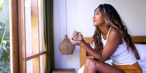 pensive young asian filipino woman holding glass of water looking out through the window sitting on bed female relaxing in the morning copy spacehydration concept