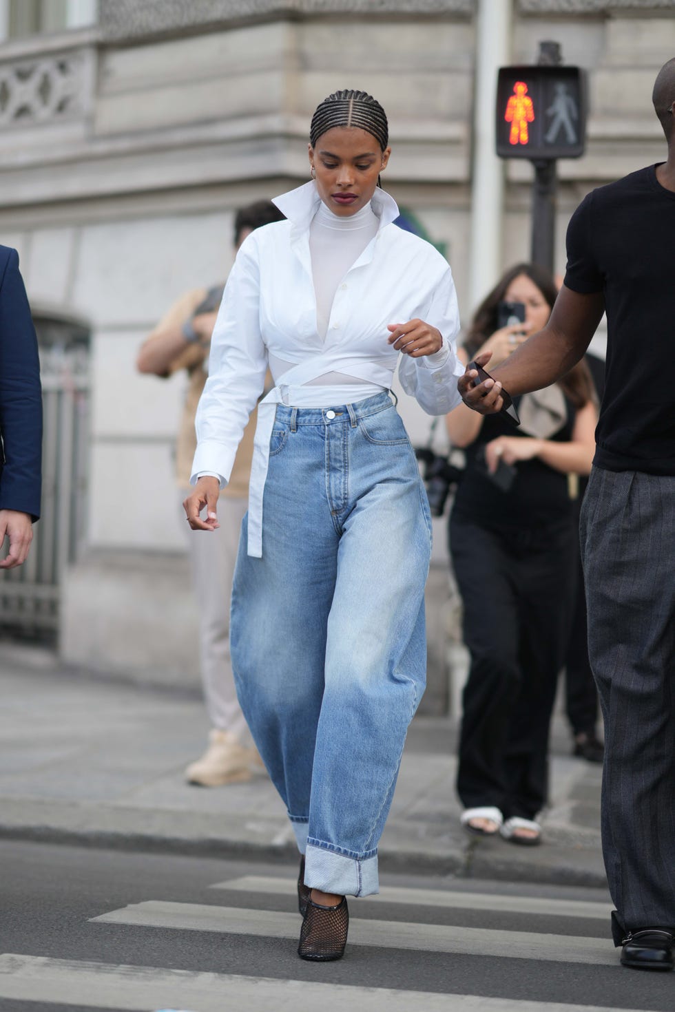 paris, france july 02: tina kunakey wears gold earrings, a white turtleneck long sleeves t shirt, a white laces waist cropped waist shirt, high waist blue faded denim puffy rolled up ankle pants, black mesh / fishnet heels pointed shoes , outside alaia, on july 02, 2023 in paris, france. (photo by edward berthelot/getty images)