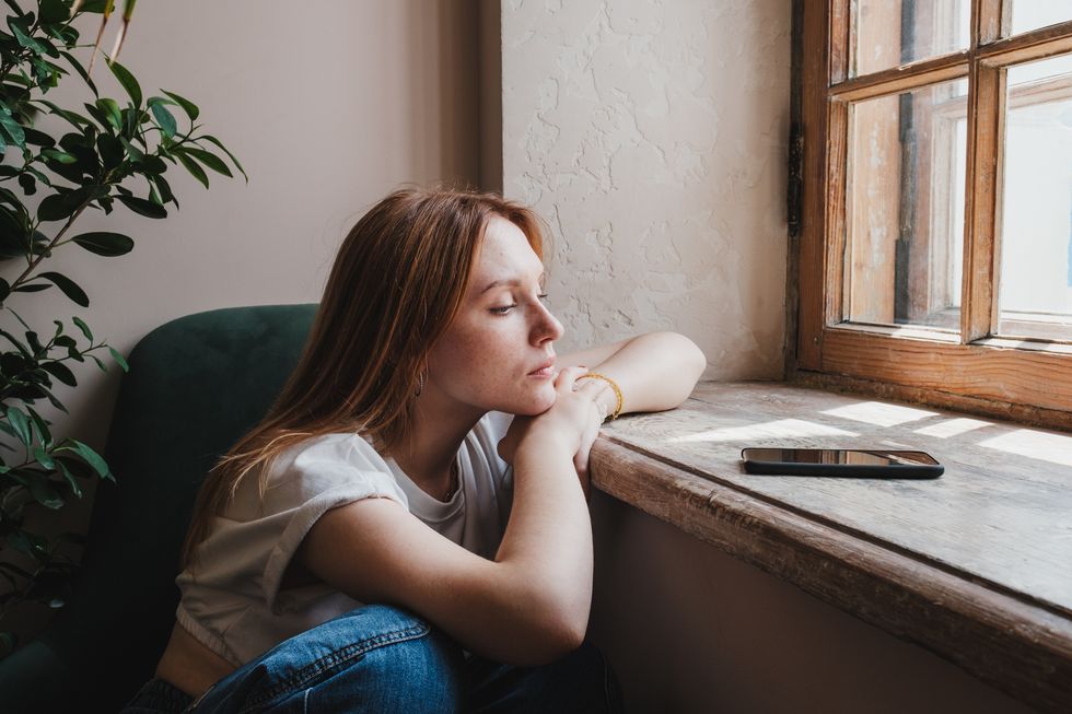 upset redhead teen girl sitting by window looking at phone waiting call from boyfriend, feeling sad and depressed teenager looking at smartphone wait for message social media depression in teens