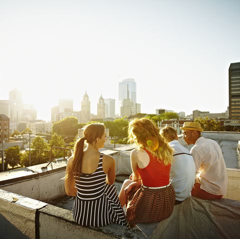 group of friends sitting on roof ledge at sunset looking out at cityscape rear view