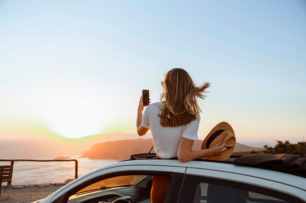 a photo of an unrecognizable woman looking at the sea and mountains from the opened roof of her car she is taking photos of a beautiful sunset