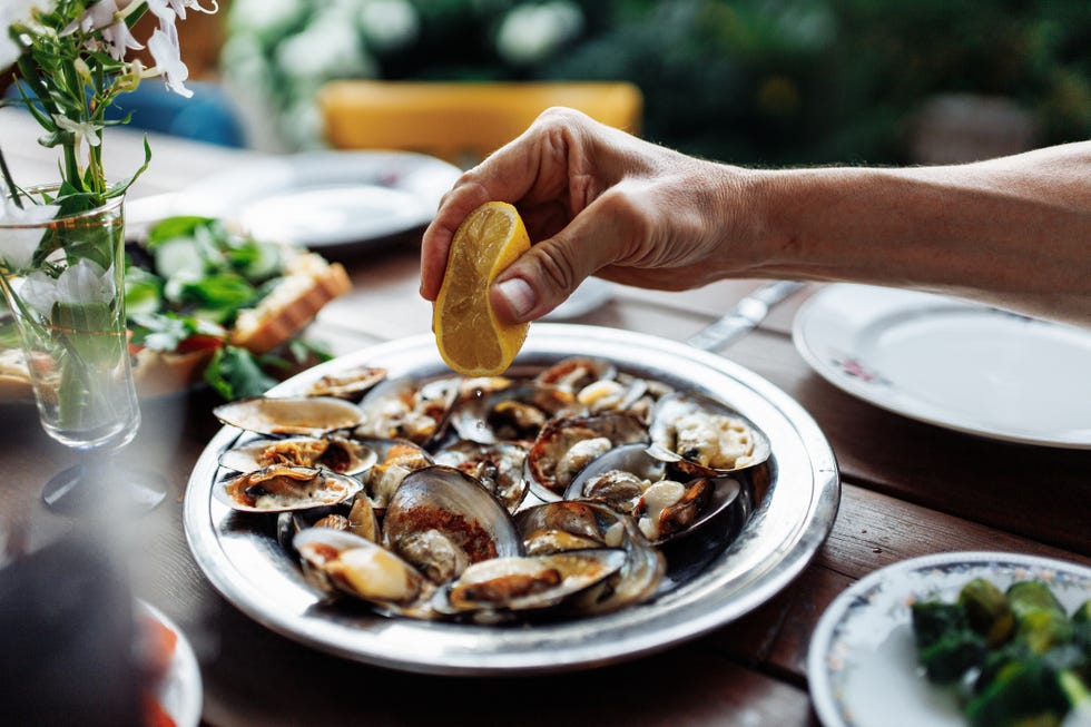 man with half of lemon in hand, adding sour taste for cooked on coals seashells by squeezing yellow fruit on mollusks. unrecognizable male seasoning and flavoring roasted oysters, having dinner at table in rustic county cottage.
