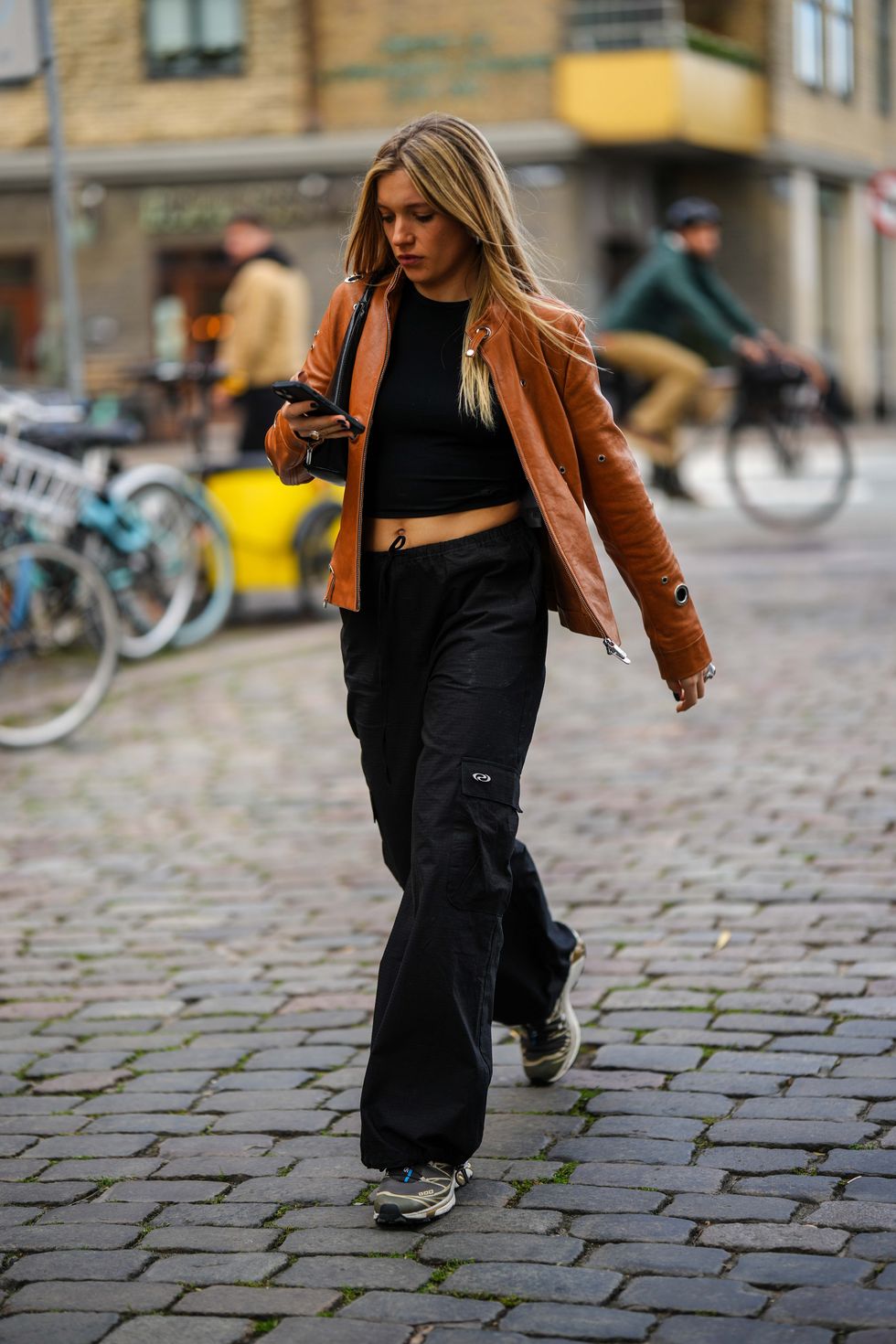 copenhagen, denmark august 08 a guest wears a black shiny leather shoulder bag, a black cropped top, a camel shiny leather zipper jacket, black cargo pants, gray sneakers from salomon , outside remain, during the copenhagen fashion week springsummer 2024 on august 08, 2023 in copenhagen, denmark photo by edward berthelotgetty images