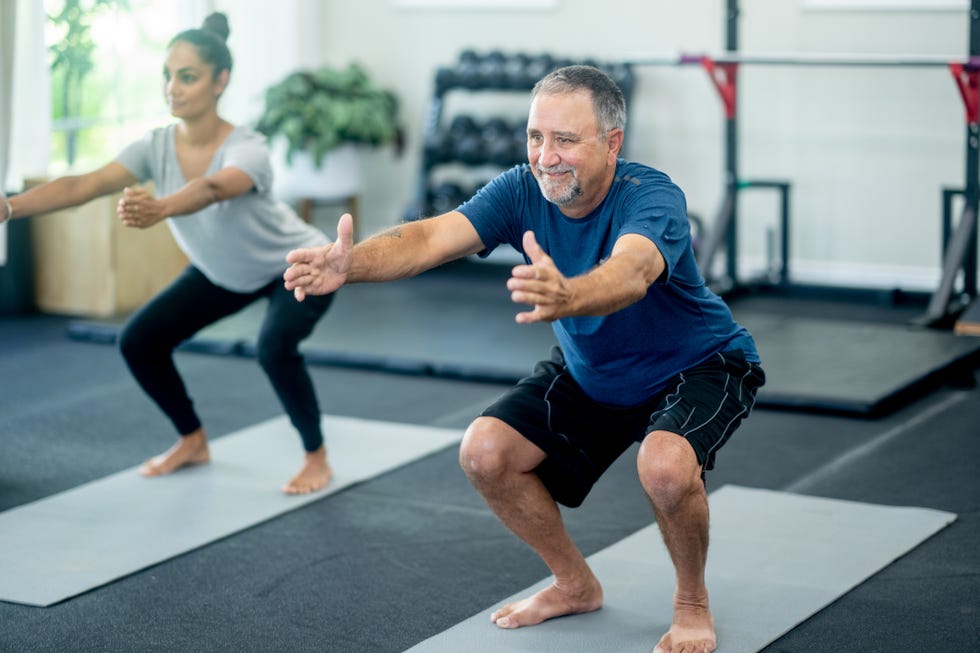 a senior gentleman stretches with his daughter in a small studio as they work to stay fit and healthy both are wearing fitness attire and are focused on holding their air squat