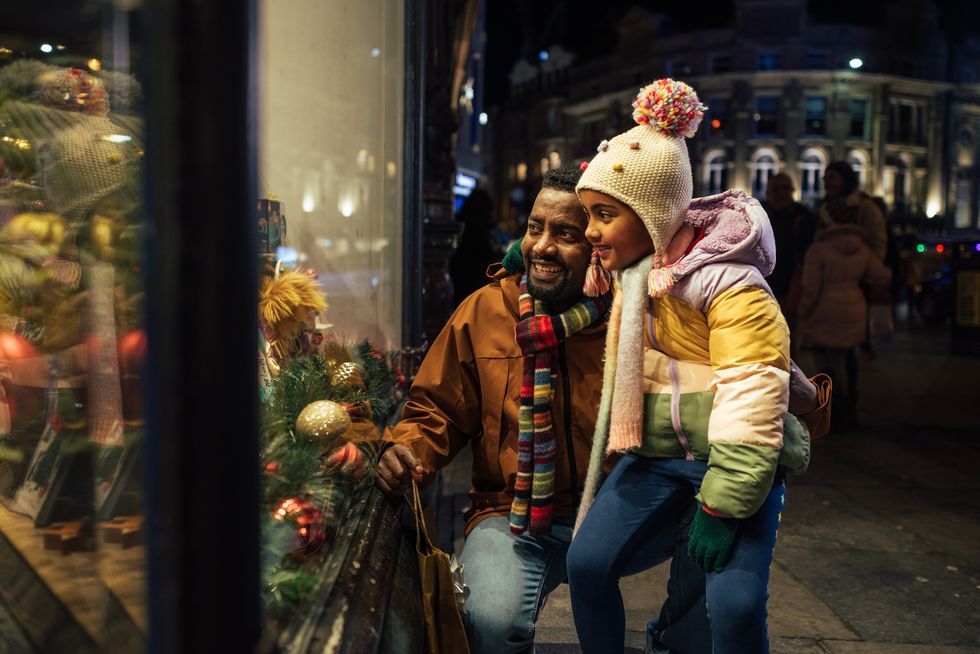 side view of a father standing outside of a store window with his young daughter, they are shopping for christmas presents and theyre wrapped up in warm clothing on a cold december night