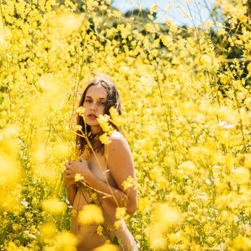 anxiety, vulnerability and acceptance fine art nude artwork of woman covered by yellow flowers in a field of yellow mustard wild flowers looking towards the camera in california