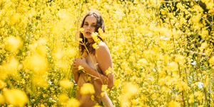 anxiety, vulnerability and acceptance fine art nude artwork of woman covered by yellow flowers in a field of yellow mustard wild flowers looking towards the camera in california
