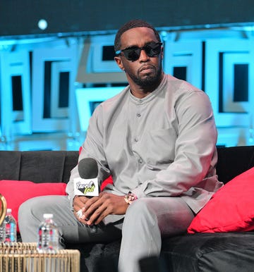 a man in a gray outfit with black sunglasses looks to the right while seated and holding a microphone at his lap