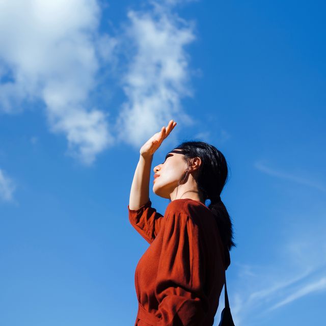 low angle portrait of young asian woman shielding eyes with her hand, standing against beautiful clear blue sky and sunlight freedom in nature connection with nature
