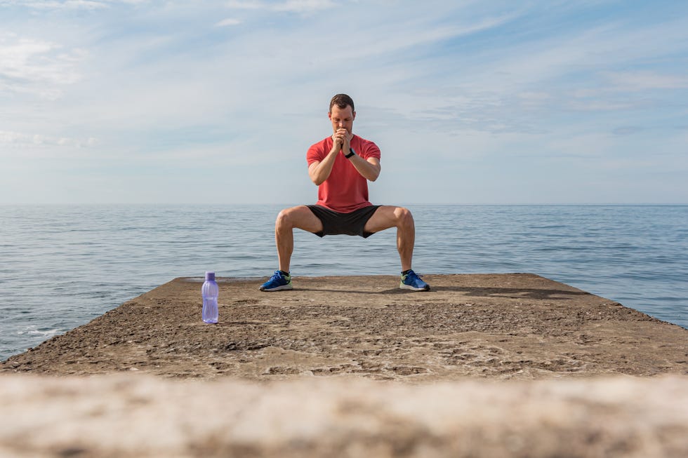 young man doing wide squatting on the beach exercise for training buttocks outdoors by the sea front view
