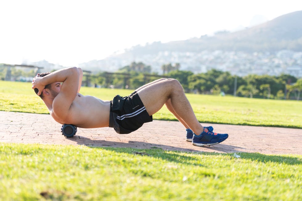 young man stretches using foam roller under his back whilst working out in a public park