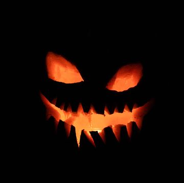 a carved pumpkin, has a flame inside it is in a dark room, so all that can be seen is the illuminated, carved face the eyes and mouth are shining orange