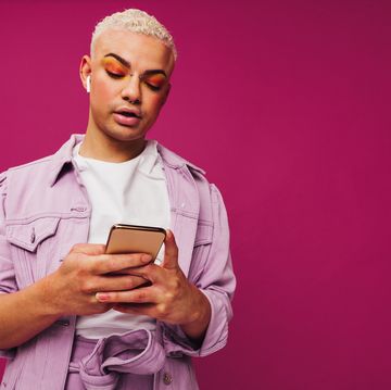 browsing trendy podcasts stylish young man using a smartphone while wearing wireless earphones in a studio non conforming queer man standing alone against a purple background