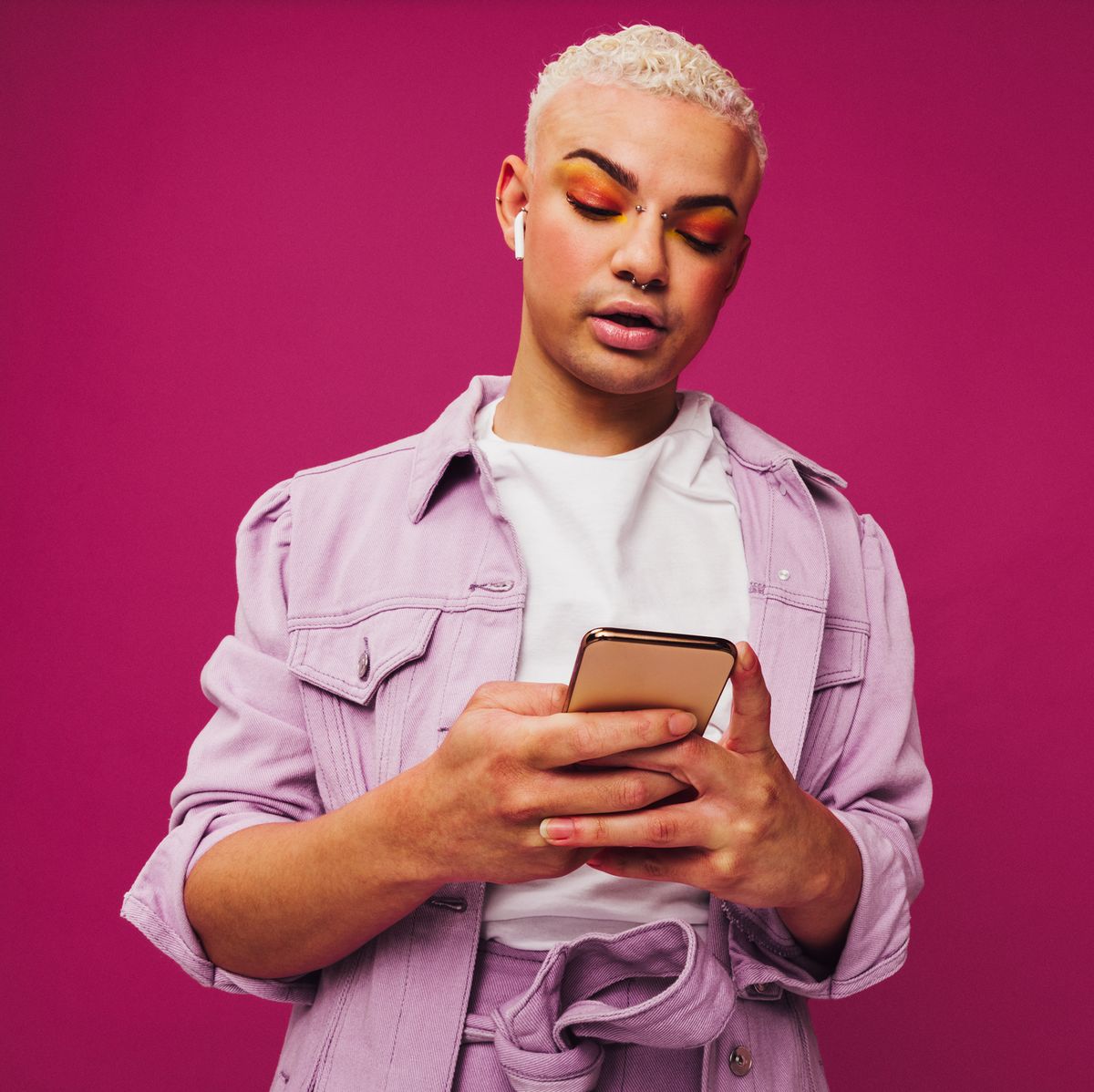 browsing trendy podcasts stylish young man using a smartphone while wearing wireless earphones in a studio non conforming queer man standing alone against a purple background
