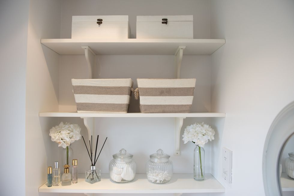 a general view of shelves in a bathroom holding perfume bottles, storage jars holding cotton buds and pads, storage baskets and boxes and scented sticks within a home