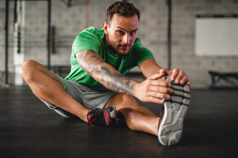 full length front view of caucasian man in late 20s looking at camera while sitting on gym floor and stretching hamstrings