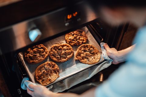 over the shoulder view of young woman taking out freshly baked cookies from the oven