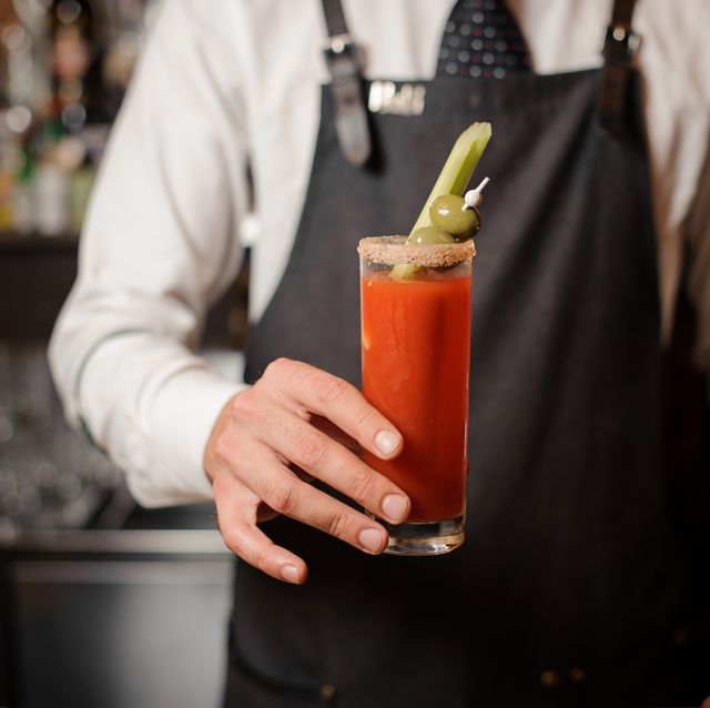 bartender in the white shirt and dark apron holding in hand a bloody mary cocktail with two olives and celery on the bar counter
