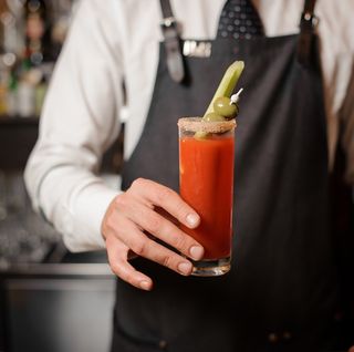 bartender in the white shirt and dark apron holding in hand a bloody mary cocktail with two olives and celery on the bar counter