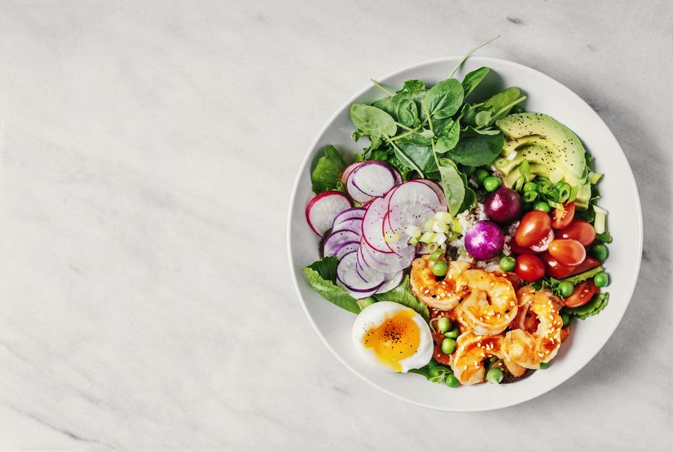 healthy lunch bowl with greens, avocado, cherry tomatoes, radish, boiled egg, and shrimp on white background