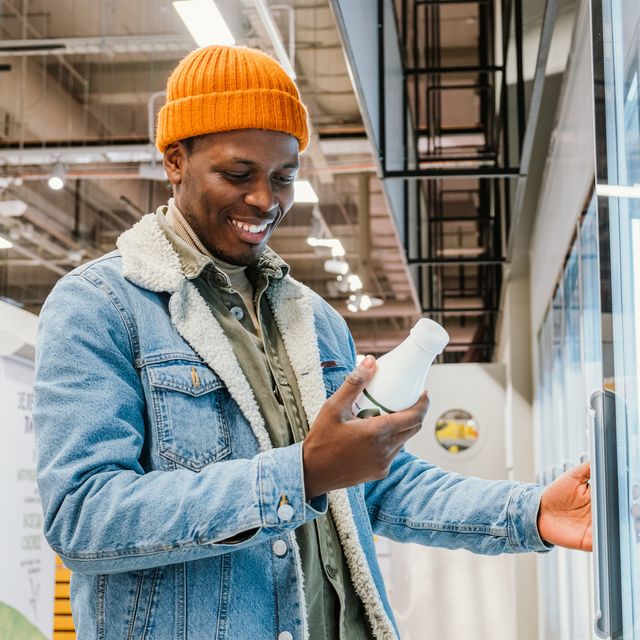 satisfied african millennial man in an orange hat in a supermarket chooses bottled milk standing at the refrigerator, and reads the composition of the product, smiles found the right product
