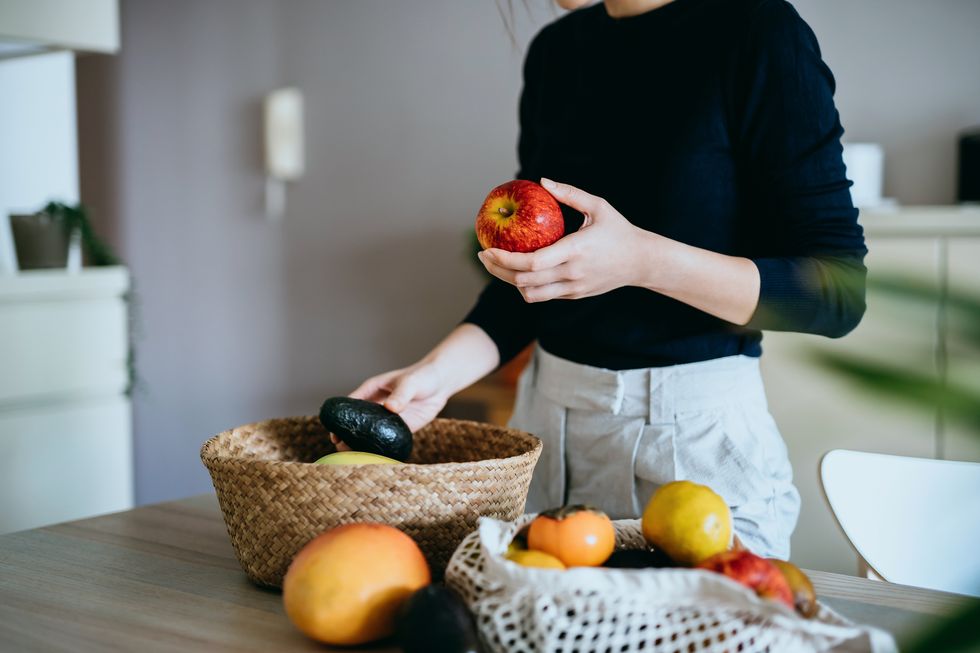 cropped shot of young asian woman coming home from grocery shopping, unpacking and organizing fresh and healthy organic fruits and veggies from a reusable bag to a rattan basket on the table responsible shopping, zero waste, sustainable lifestyle concept