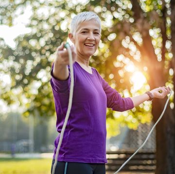 happy woman in purple shirt exercising with jump rope in a park in front of a tree