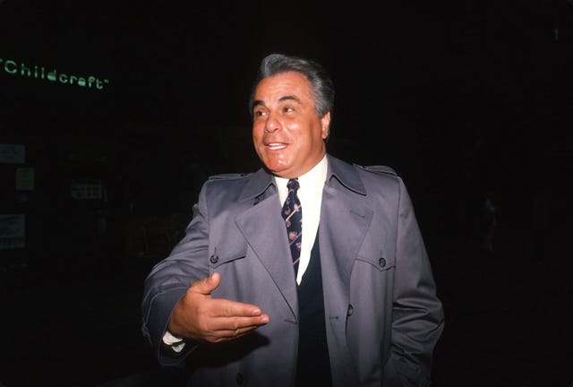 john gotti stands in a gray coat and suit in darkness, he gestures one hand in front of his chest and smiles as he looks left