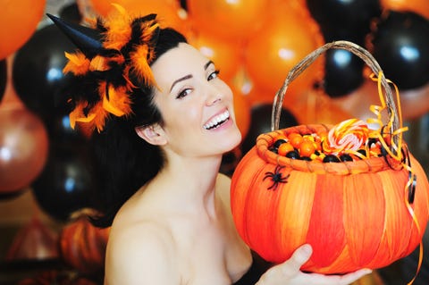 beautiful smiling brunette woman dressed up for halloween with a feather hat, holding an orange basket filled with candies, with black and orange balloons in the background