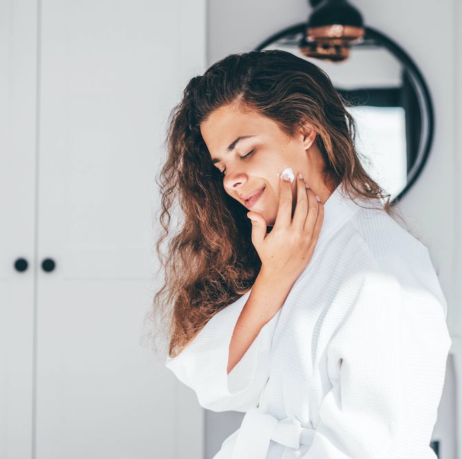 young woman with long curly hair in bathrobe enjoys daily routine applying moisturizing cream on face in bedroom at home closeup mirror reflection
