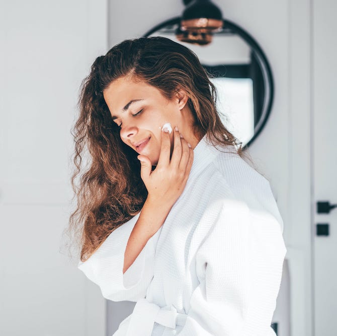 young woman with long curly hair in bathrobe enjoys daily routine applying moisturizing cream on face in bedroom at home closeup mirror reflection