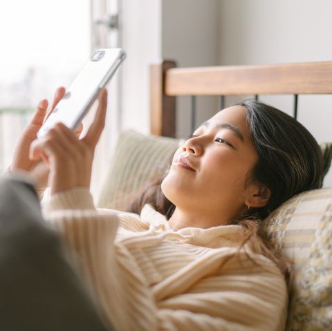 a young and beautiful woman is lying in bed and using a smart phone happily in the morning