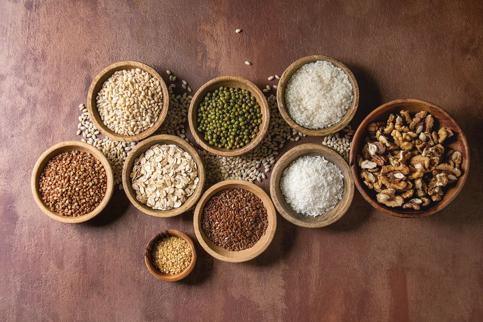 variety of raw uncooked grains superfood cereal sesame, mung bean, walnuts, tapioca, wheat, buckwheat, oatmeal, coconut, rice in wooden bowls over brown background flat lay, space