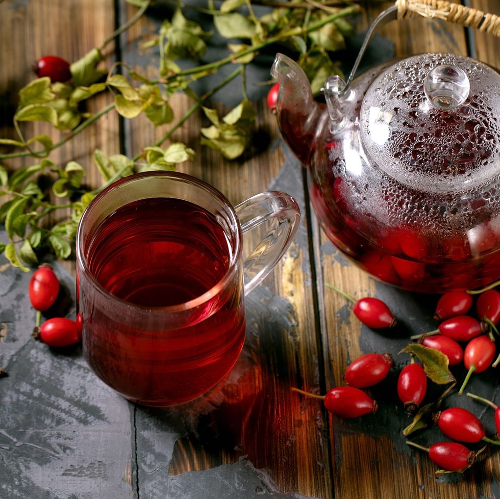cup of rose hip berries herbal hot tea and glass teapot standing on old wooden plank table with wild autumn berries around winter vitamin beverage