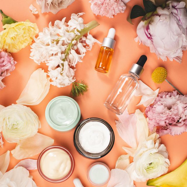 top view of jars with creams and bottles with essential oils placed amidst various fresh flowers on bright orange background