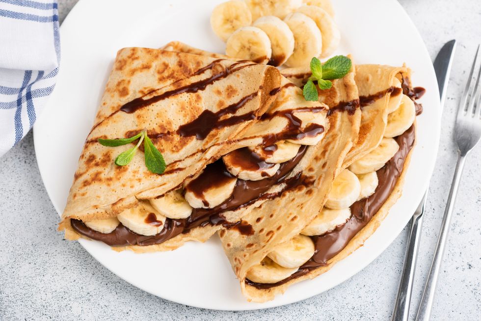 crepes or blini stuffed with chocolate hazelnut spread, banana on a white plate closeup view
