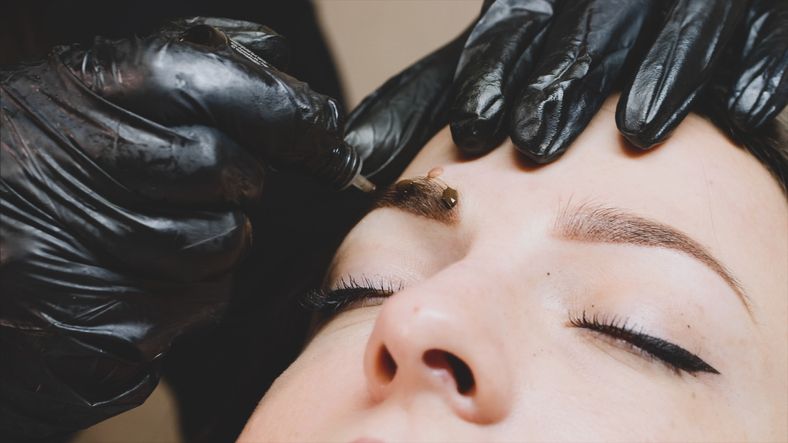 the master in black gloves is adding some drops of the brown pigment on the clients eyebrow after the microblading procedure to make a mask above the brow close up